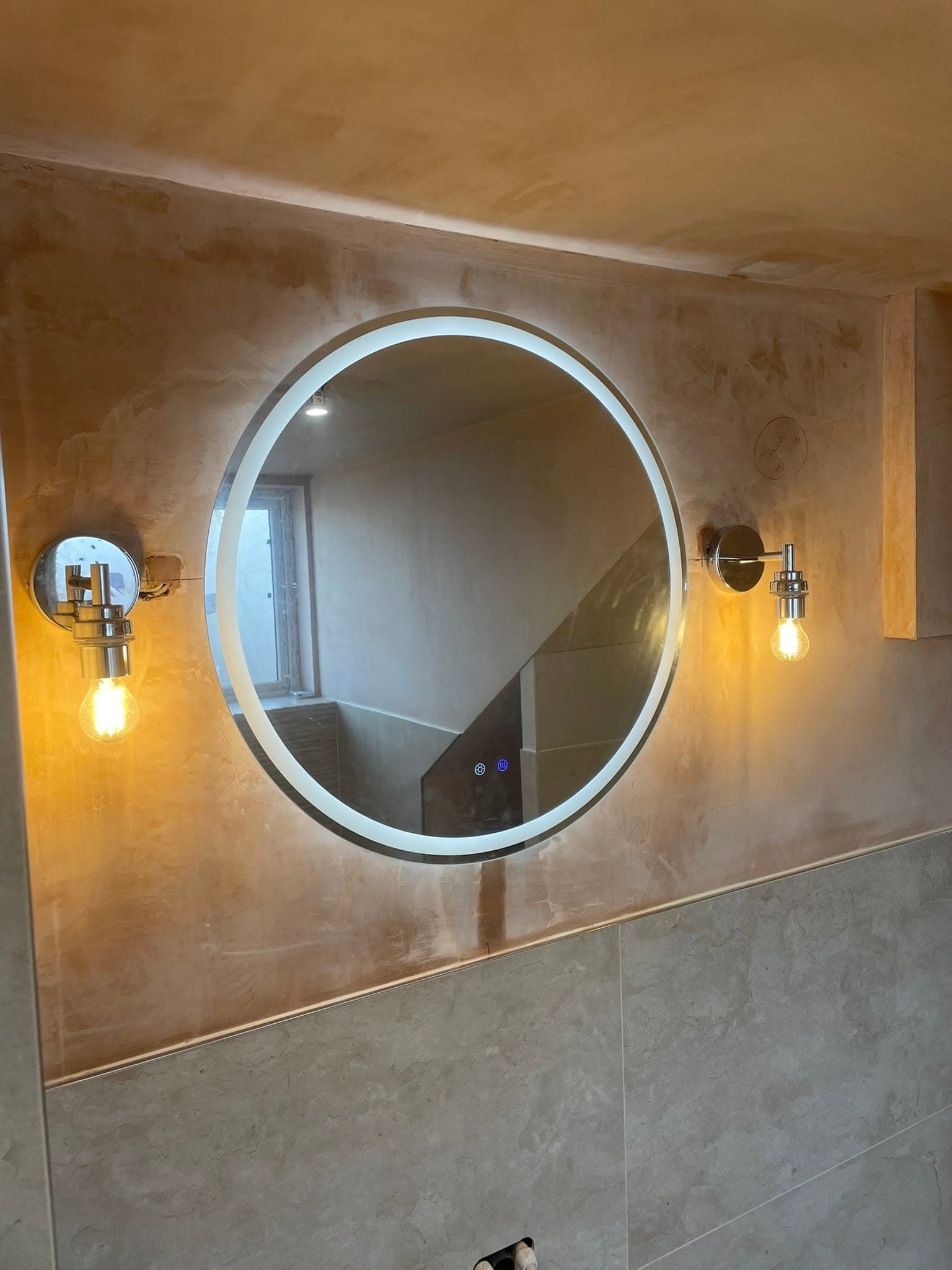 Luxury bathroom with a large LED backlit circular mirror flanked by chrome wall sconces on venetian plaster
