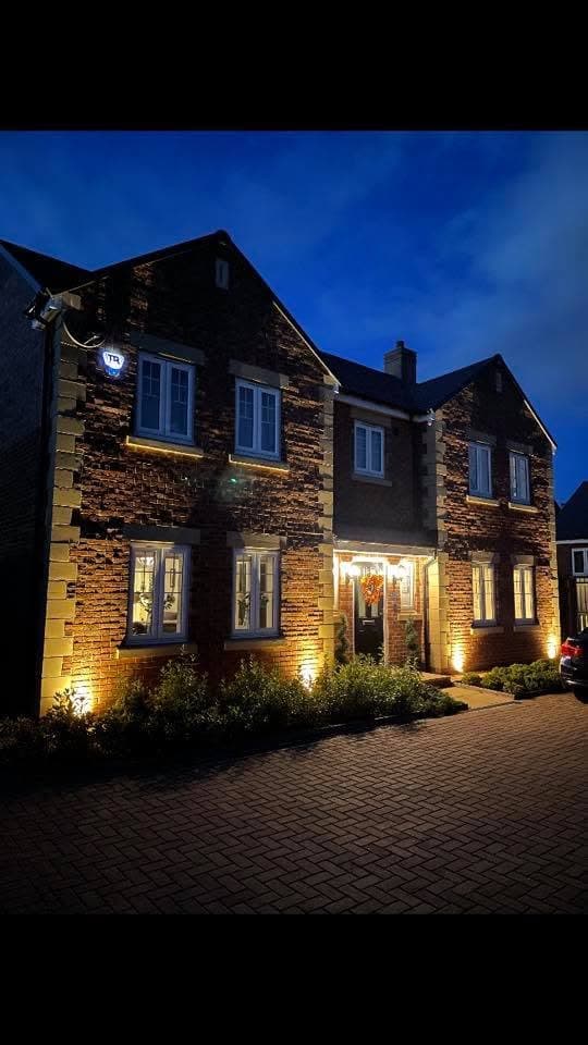 Stone detached home lit at night with warm uplights, security camera and feature front-door lighting